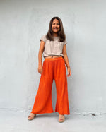 Woman wearing orange pants and a beige top against a white wall