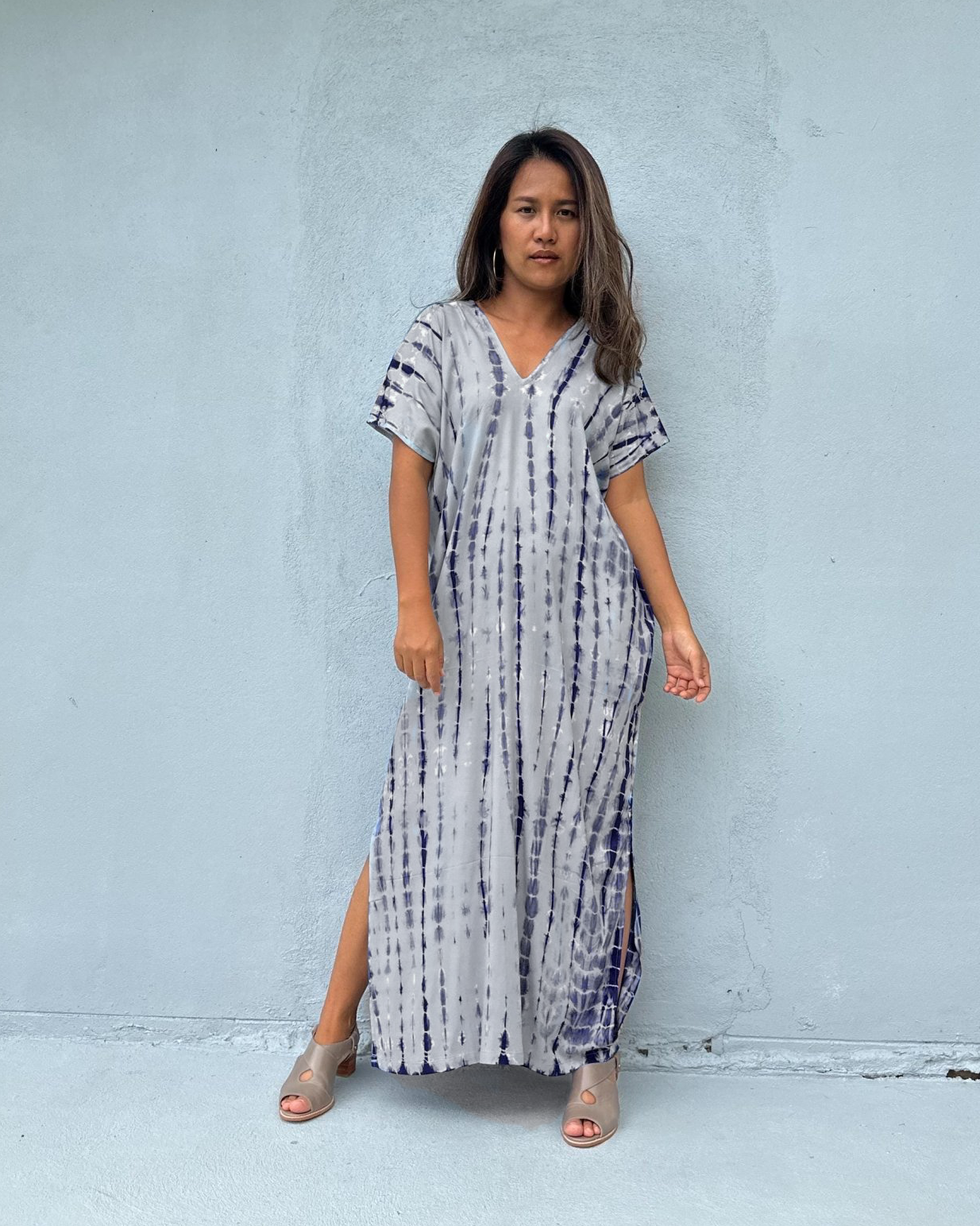 A woman stands against a light gray concrete wall wearing a loose, short-sleeve maxi dress with a white base and vertical tie-dye style blue and purple stripes. She is wearing clear high-heel sandals.