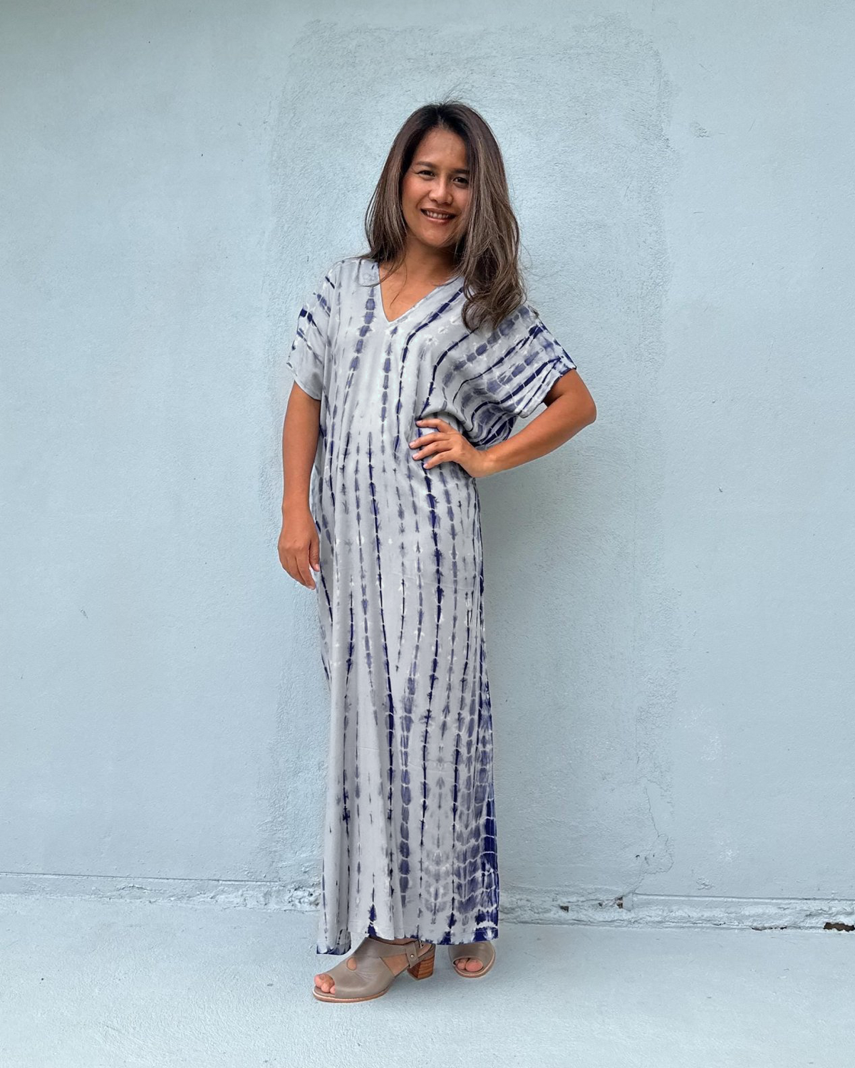 A woman modeling a long, loose-fitting maxi dress with a vertical tie-dye pattern in navy blue and white, standing against a light gray concrete wall.