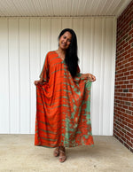 Woman wearing an orange and green patterned dress standing against a white wall with a brick column.
