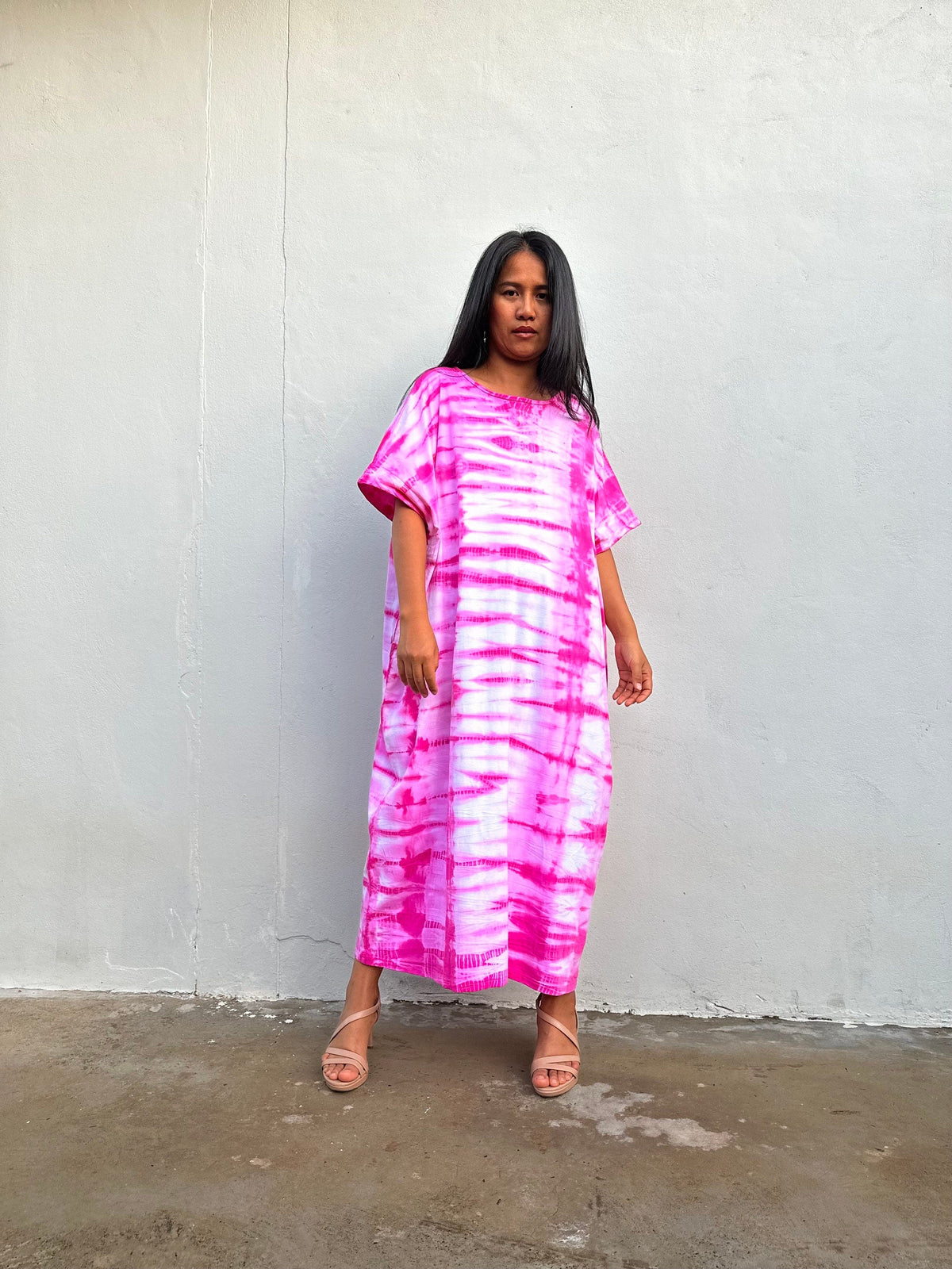 Woman wearing a pink tie-dye dress against a white wall.