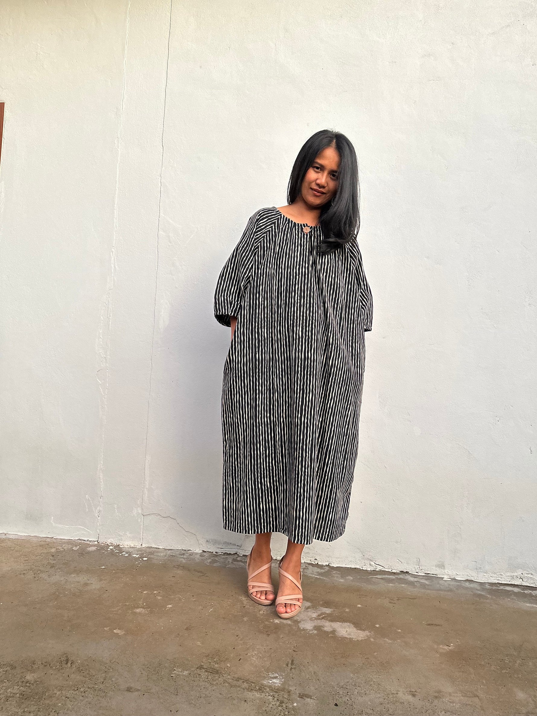 A woman modeling a loose-fitting, midi-length dress with a black and white vertical stripe pattern, standing against a textured off-white wall.