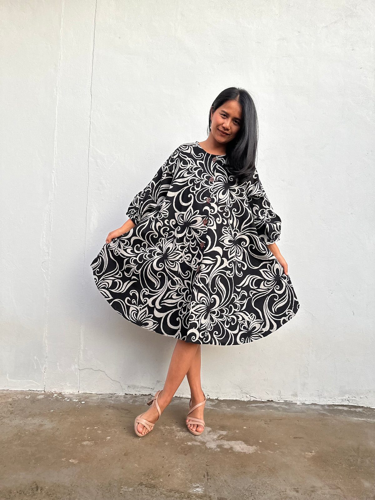 A woman modeling a black and white floral print dress with long sleeves, standing against a white textured wall.