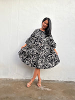 A woman modeling a black and white floral print dress with long sleeves, standing against a white textured wall.