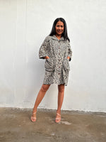A woman modeling a short-sleeve button-up shirt dress with a black and white abstract animal print, standing against a white wall.