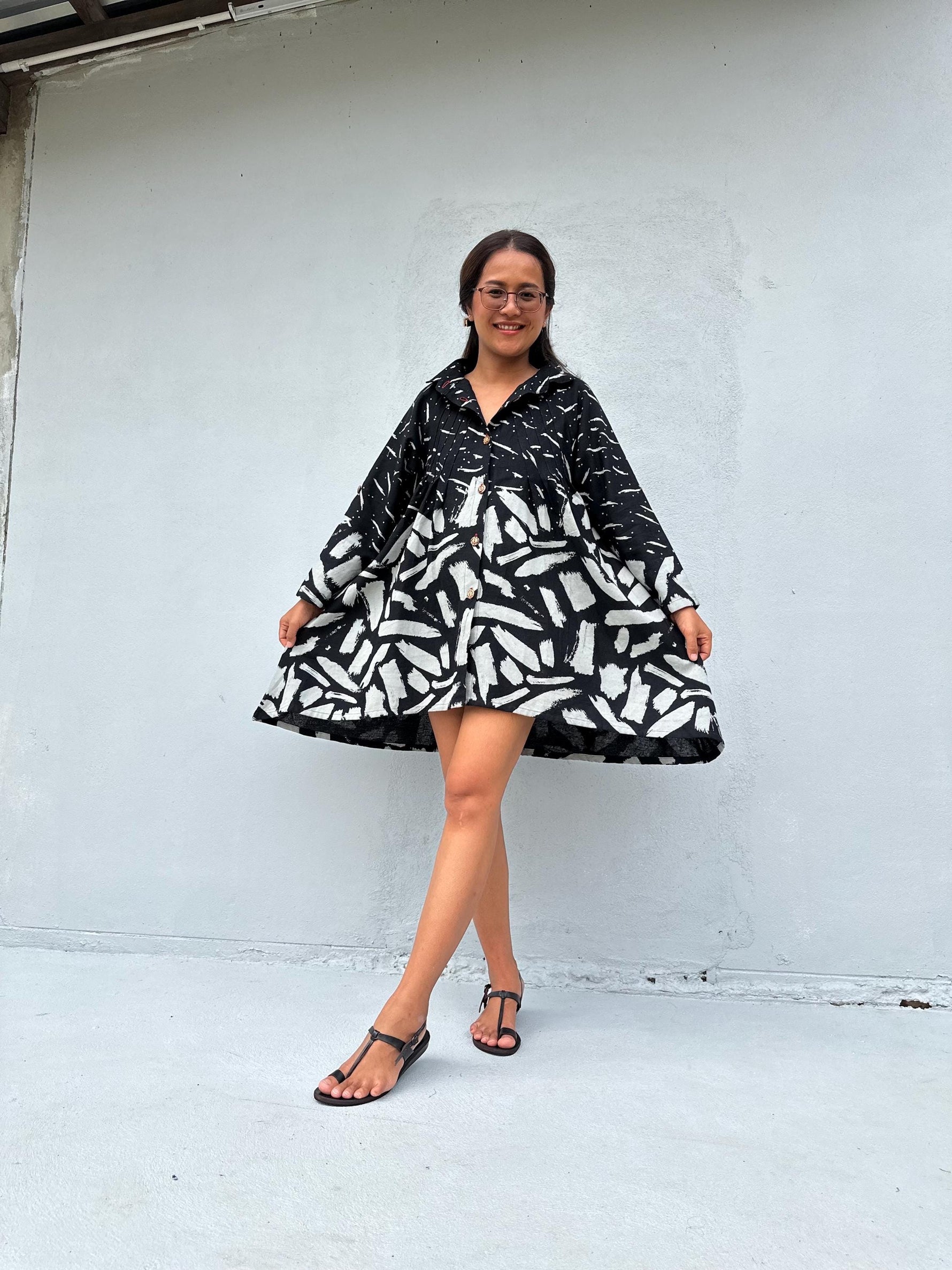 A woman modeling a black and white abstract patterned mini dress with long sleeves and a button-up front, standing against a light gray concrete wall.