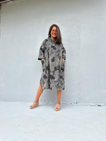 A woman wearing a gray and black snakeskin-patterned midi dress with pockets, standing against a light gray concrete wall.