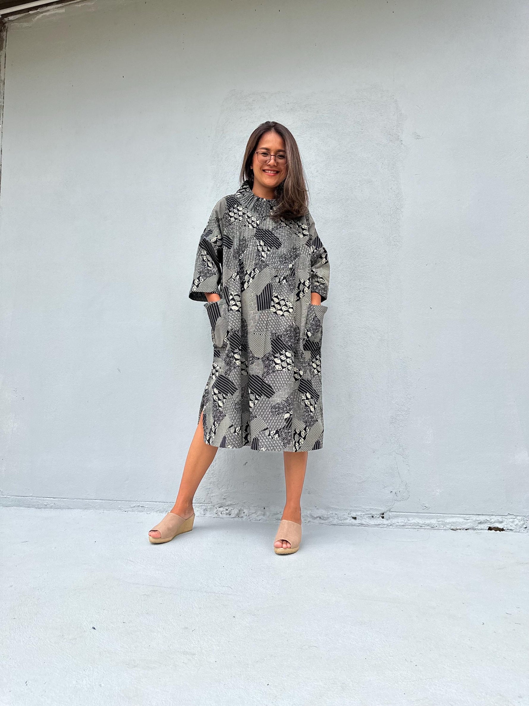 A woman wearing a gray and black snakeskin-patterned midi dress with pockets, standing against a light gray concrete wall.