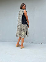 A woman wearing a sleeveless black and beige abstract patterned vest over a black top, paired with beige wedge sandals, standing against a light gray concrete wall.