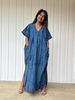 Woman wearing a blue tie-dye dress standing against a white paneled wall.