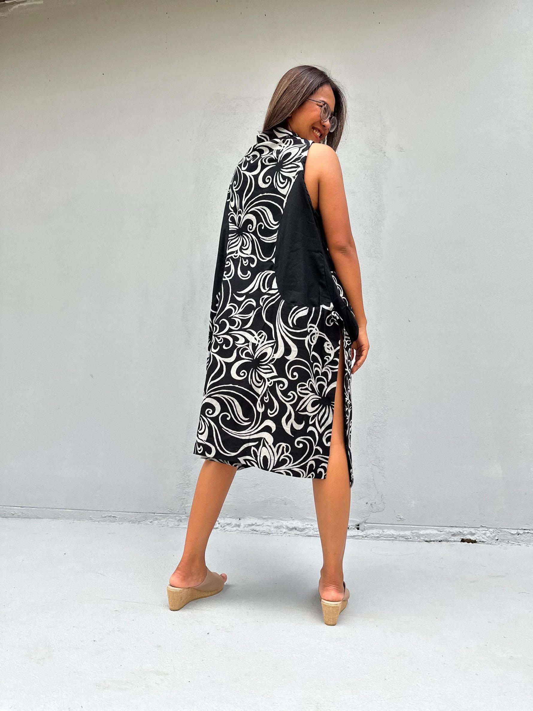 A woman wearing a sleeveless black and white patterned midi dress with a high collar, photographed from behind against a plain light gray wall.