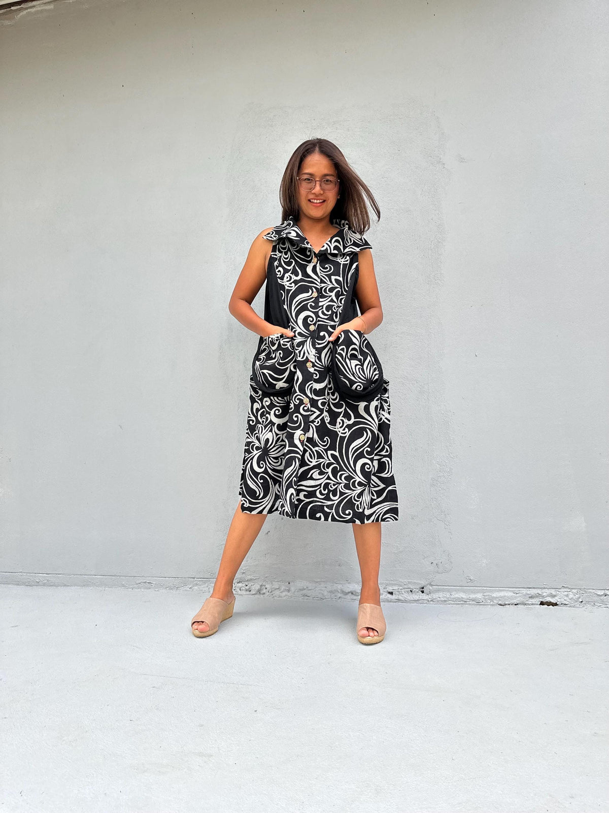 A woman modeling a sleeveless midi dress with a black and white swirling damask pattern, standing against a light gray concrete wall.