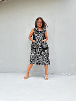 A woman modeling a sleeveless midi dress with a black and white swirling damask pattern, standing against a light gray concrete wall.