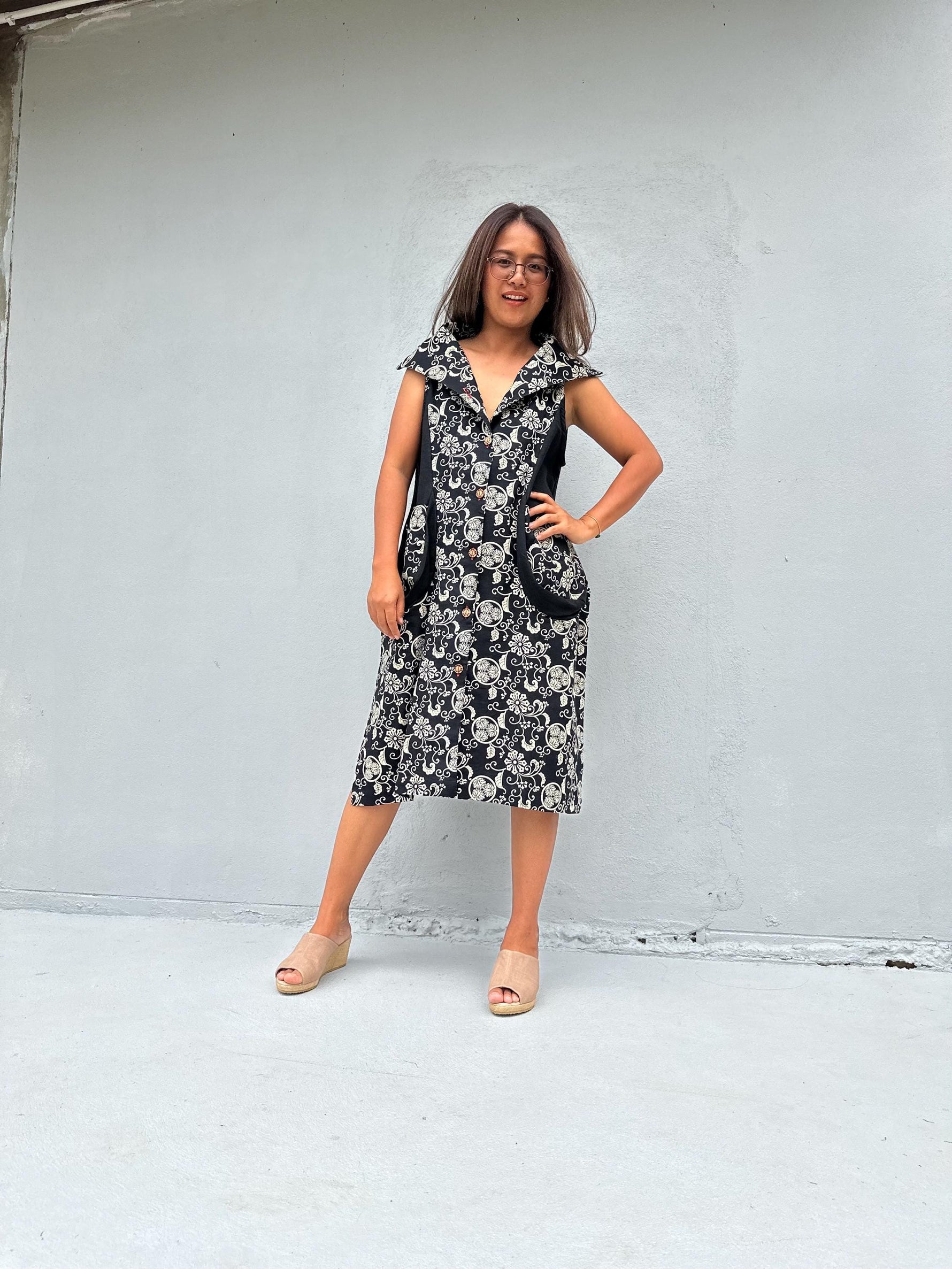 A woman wearing a black and white floral print midi dress with a V-neckline and side pockets, standing against a light gray concrete wall.