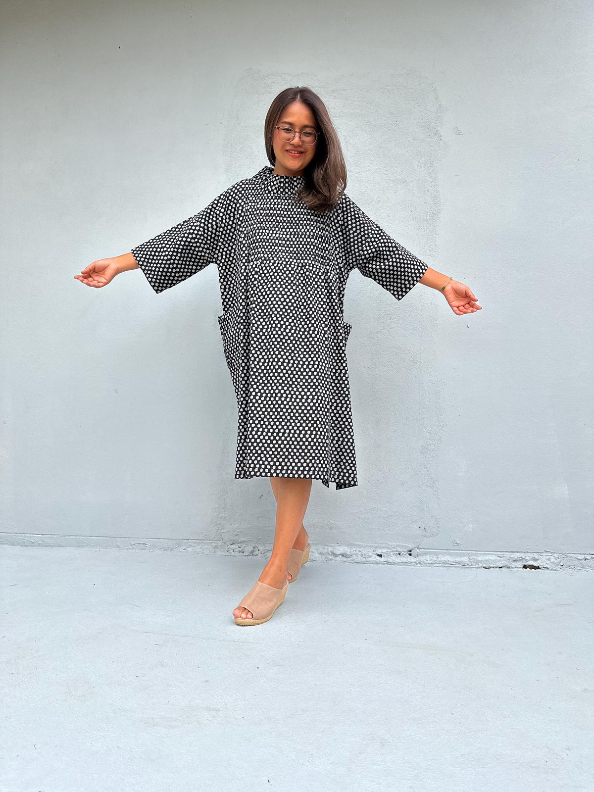 A woman modeling a loose-fitting black and white patterned dress with wide three-quarter sleeves and side pockets, standing against a light gray concrete wall with her arms outstretched.