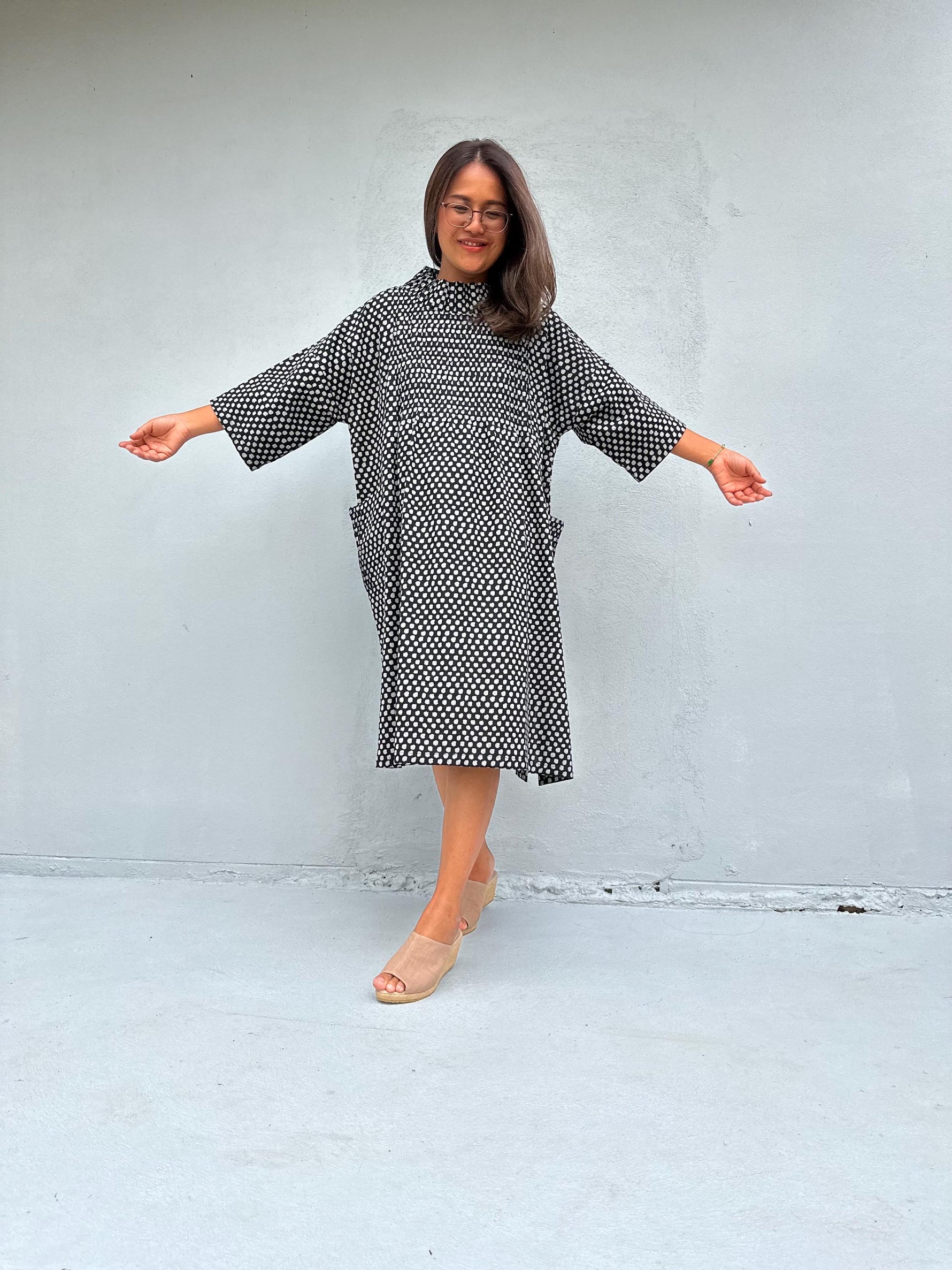 A woman modeling a loose-fitting black and white patterned dress with wide three-quarter sleeves and side pockets, standing against a light gray concrete wall with her arms outstretched.