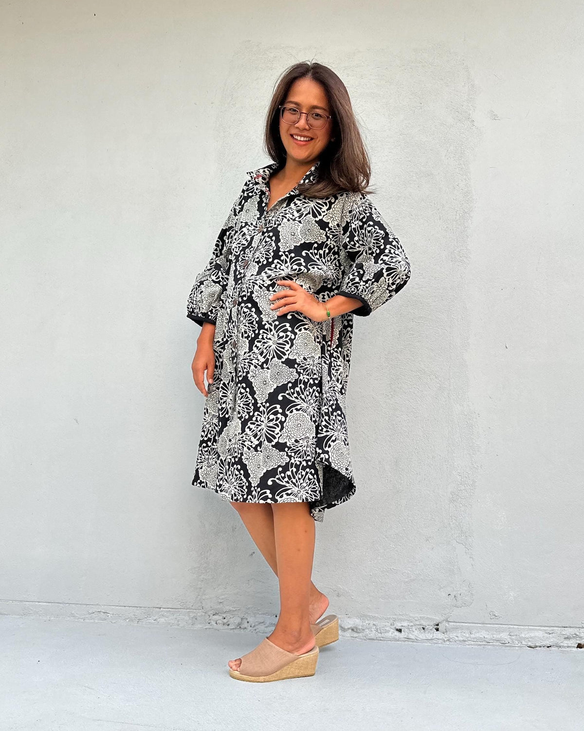 A woman wearing a black and white patterned long-sleeve dress and beige wedge sandals, standing against a plain light gray wall.