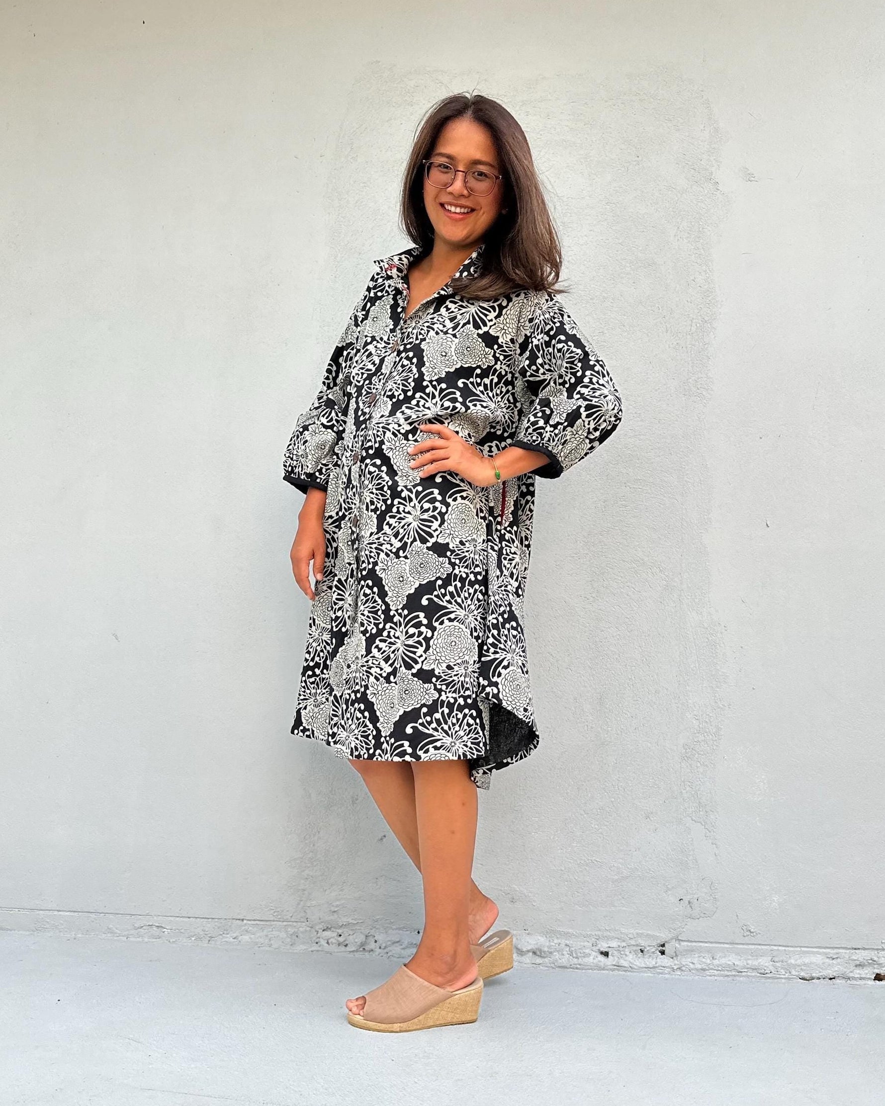 A woman wearing a black and white patterned long-sleeve dress and beige wedge sandals, standing against a plain light gray wall.