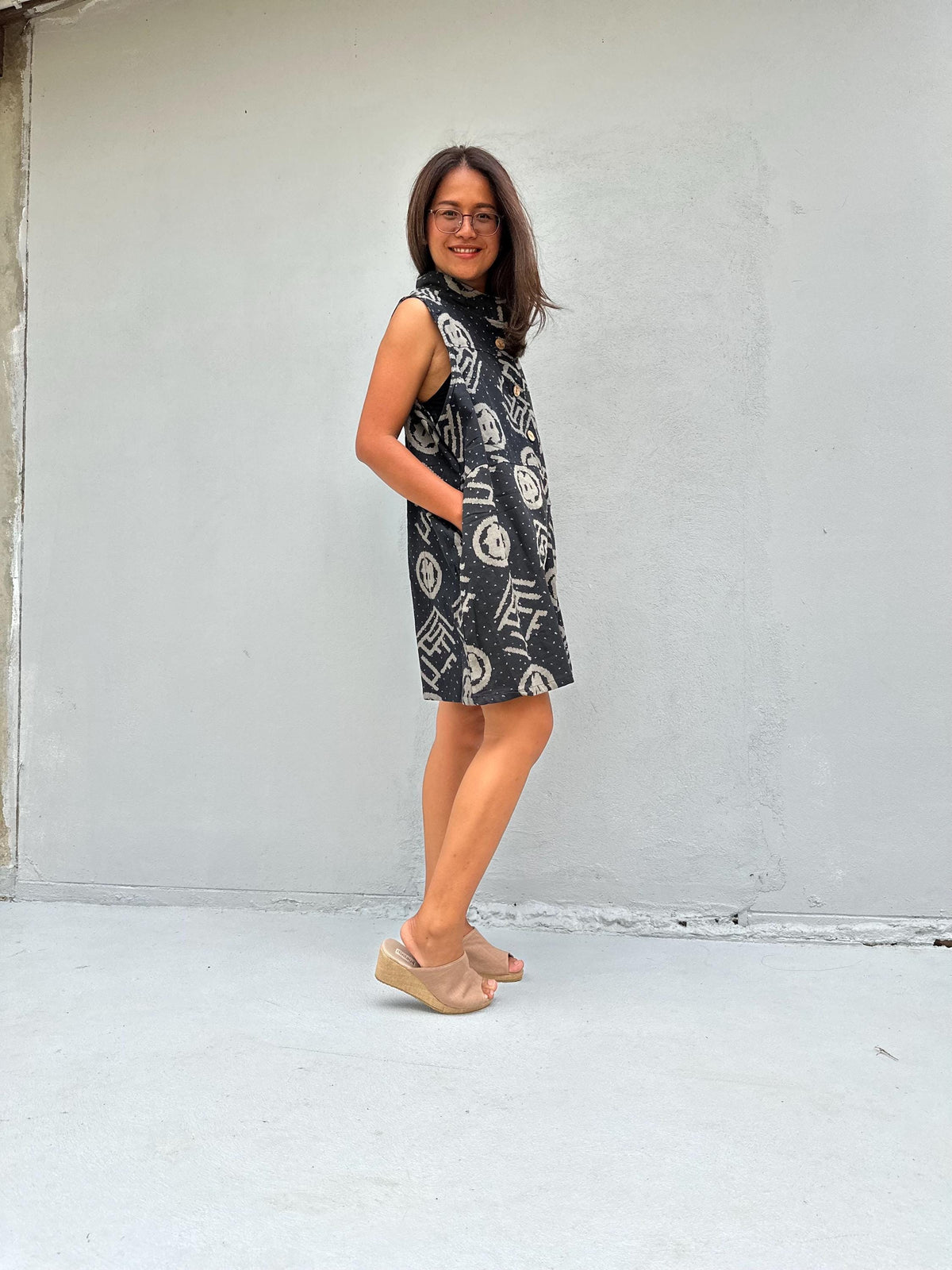 A woman wearing a sleeveless black dress with a white graffiti-style pattern and beige wedge sandals, standing against a light gray concrete wall.