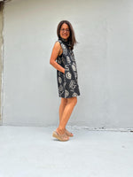 A woman wearing a sleeveless black dress with a white graffiti-style pattern and beige wedge sandals, standing against a light gray concrete wall.
