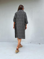 A woman viewed from behind wearing a black and white polka dot shirt dress with elbow-length sleeves, paired with tan wedge slide sandals, standing against a plain light gray wall.