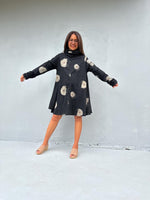 A woman modeling a black long-sleeve tunic dress with a white polka dot pattern and large white skull print, standing against a light gray concrete wall.