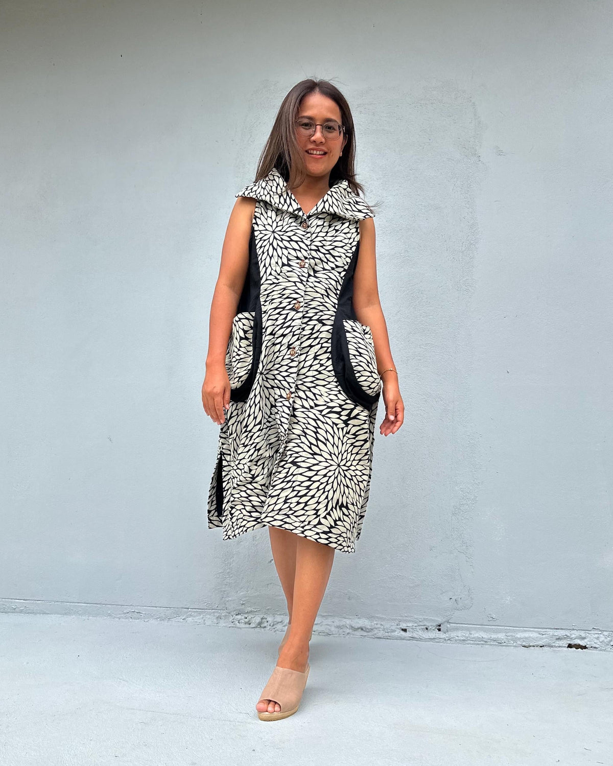 A woman modeling a sleeveless black and white patterned dress with black side pockets, standing against a light gray concrete wall.