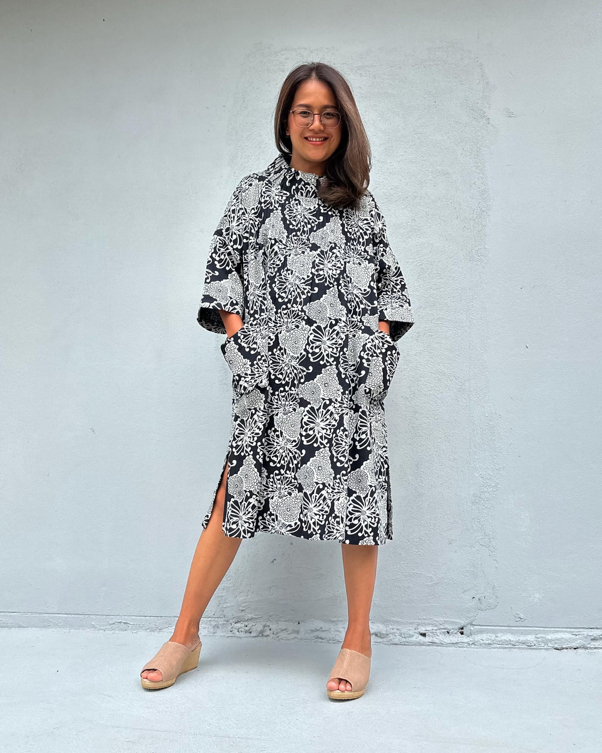 A woman wearing a black and white snakeskin print midi dress with side slits and elbow-length sleeves, standing against a light gray concrete wall.