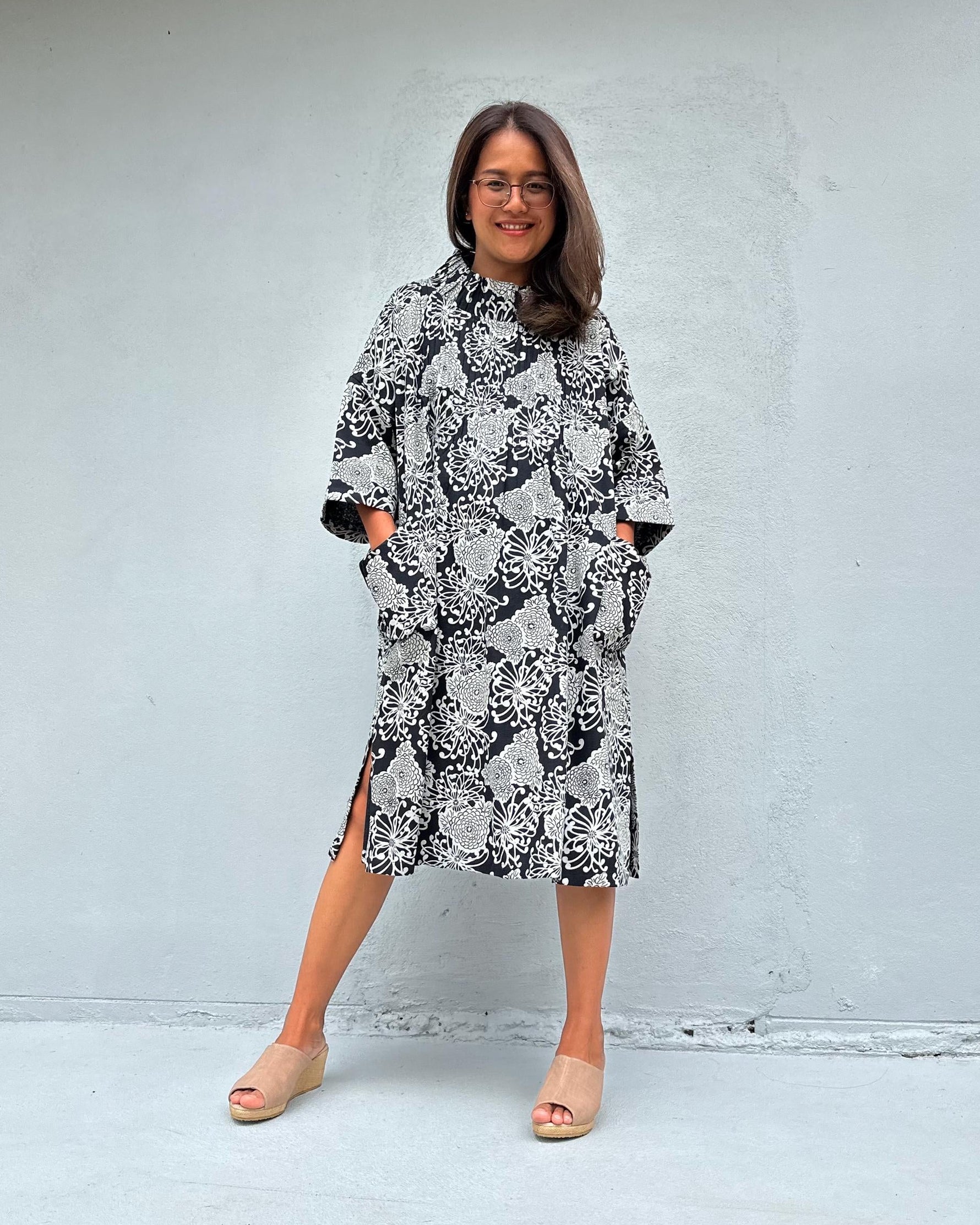 A woman wearing a black and white snakeskin print midi dress with side slits and elbow-length sleeves, standing against a light gray concrete wall.