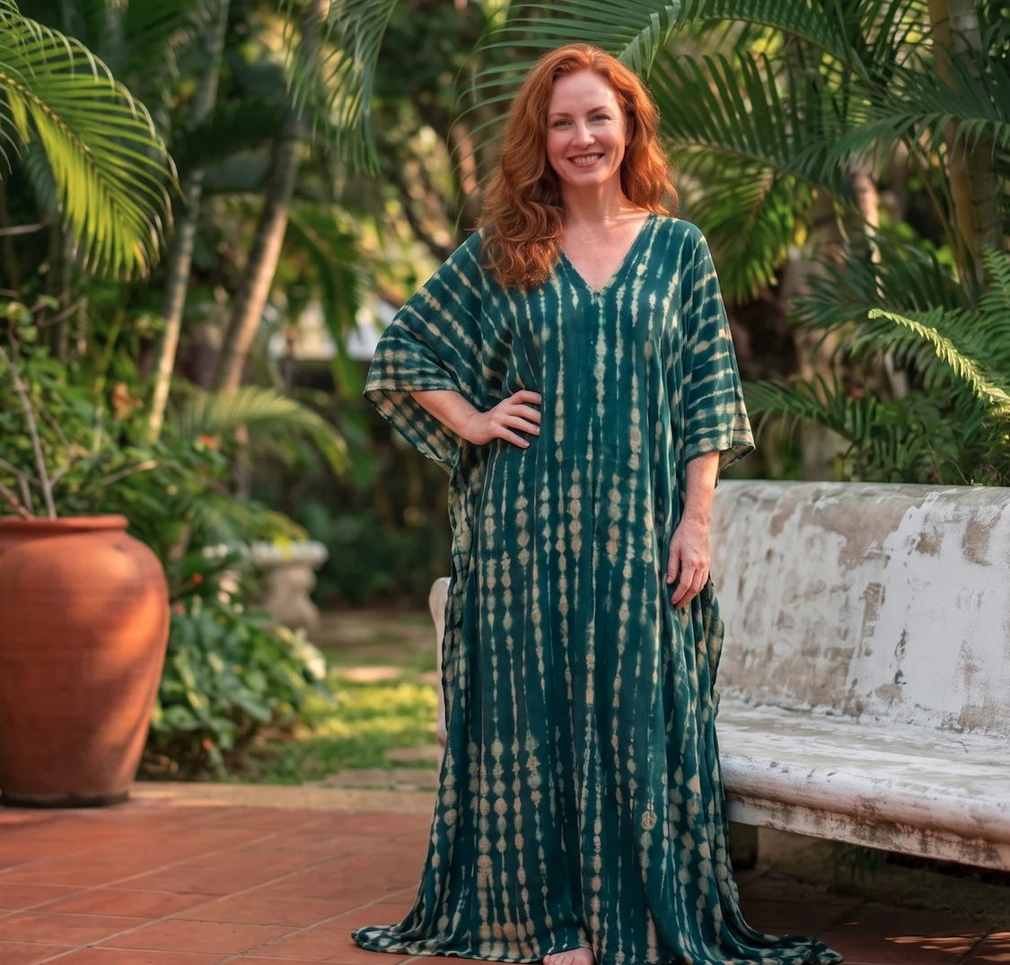 A smiling woman with red hair stands outdoors in a lush tropical garden wearing a long, flowing tall kaftan dress with a green tie-dye pattern. She is positioned in front of a weathered white bench and tropical foliage.