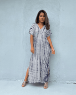 A woman stands against a light gray concrete wall wearing a loose, short-sleeve maxi dress with a white base and vertical tie-dye style blue and purple stripes. She is wearing clear high-heel sandals.