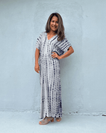 A woman modeling a long, loose-fitting maxi dress with a vertical tie-dye pattern in navy blue and white, standing against a light gray concrete wall.