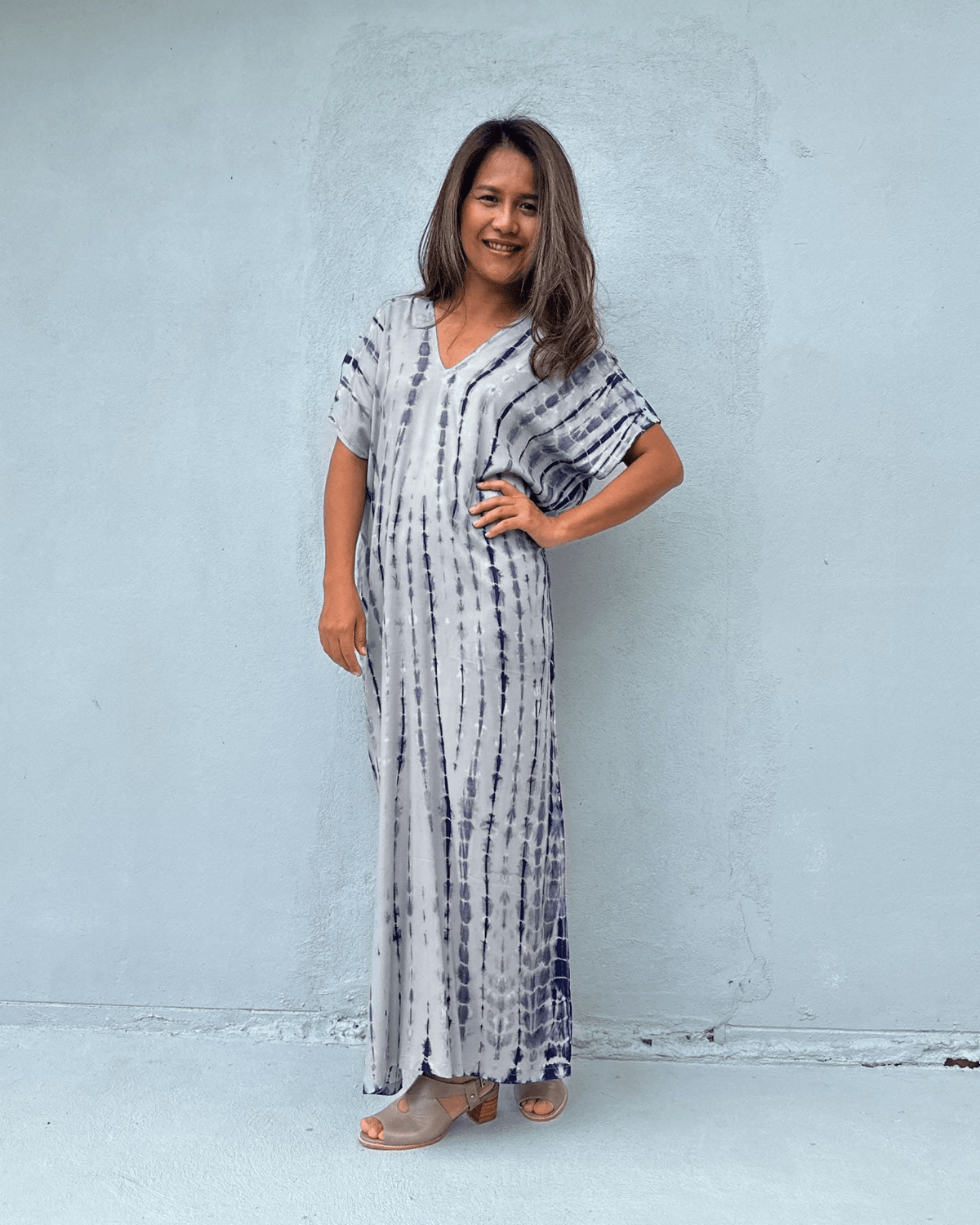 A woman modeling a long, loose-fitting maxi dress with a vertical tie-dye pattern in navy blue and white, standing against a light gray concrete wall.