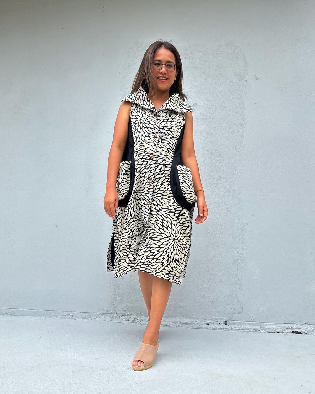 A woman modeling a sleeveless black and white patterned dress with black side pockets, standing against a light gray concrete wall.
