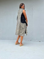 A woman wearing a sleeveless black and beige abstract patterned vest over a black top, paired with beige wedge sandals, standing against a light gray concrete wall.