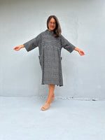 A woman modeling a loose-fitting black and white patterned dress with wide three-quarter sleeves and side pockets, standing against a light gray concrete wall with her arms outstretched.