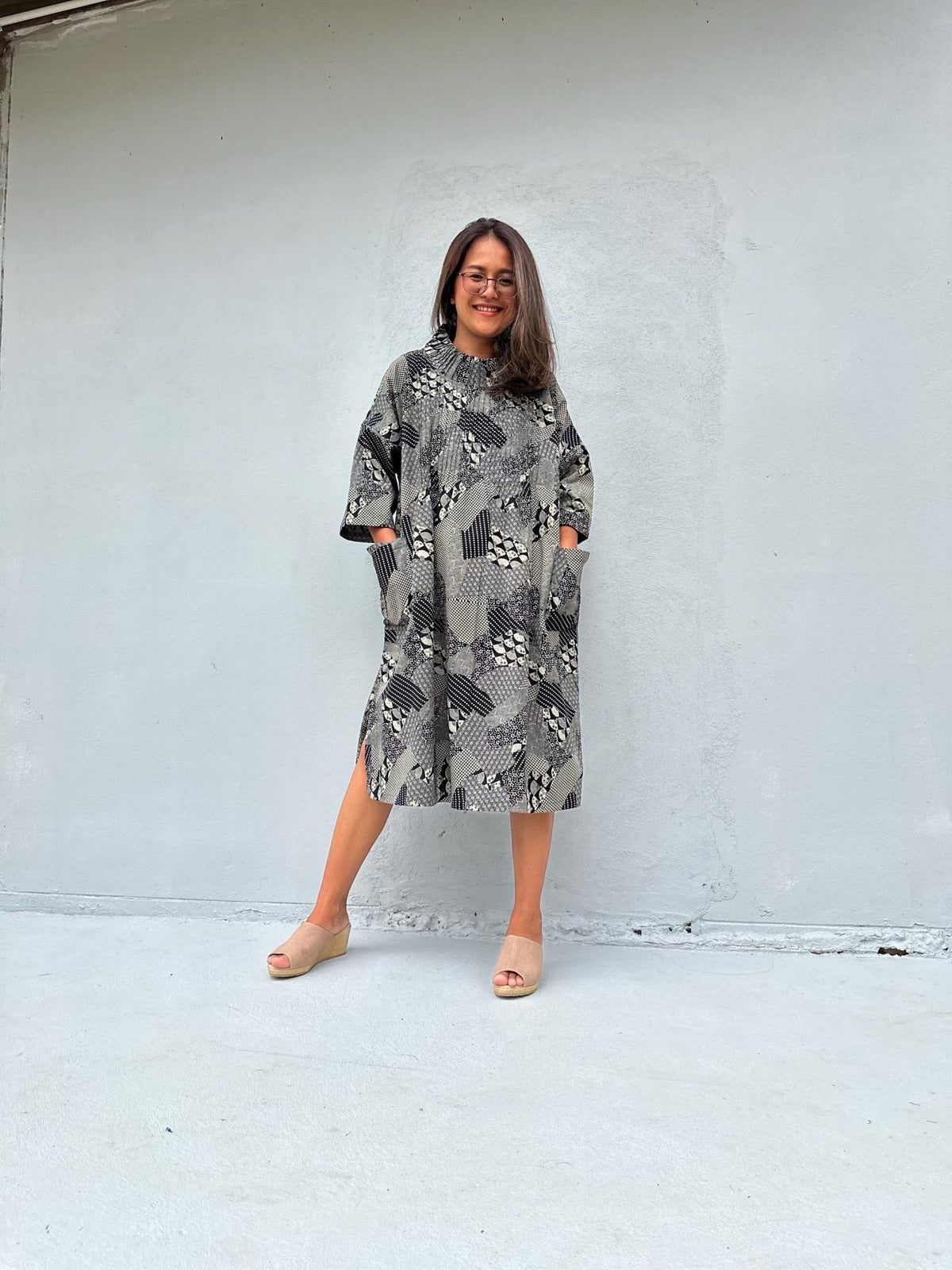 A woman wearing a gray and black snakeskin-patterned midi dress with pockets, standing against a light gray concrete wall.