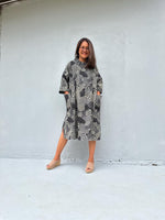 A woman wearing a gray and black snakeskin-patterned midi dress with pockets, standing against a light gray concrete wall.