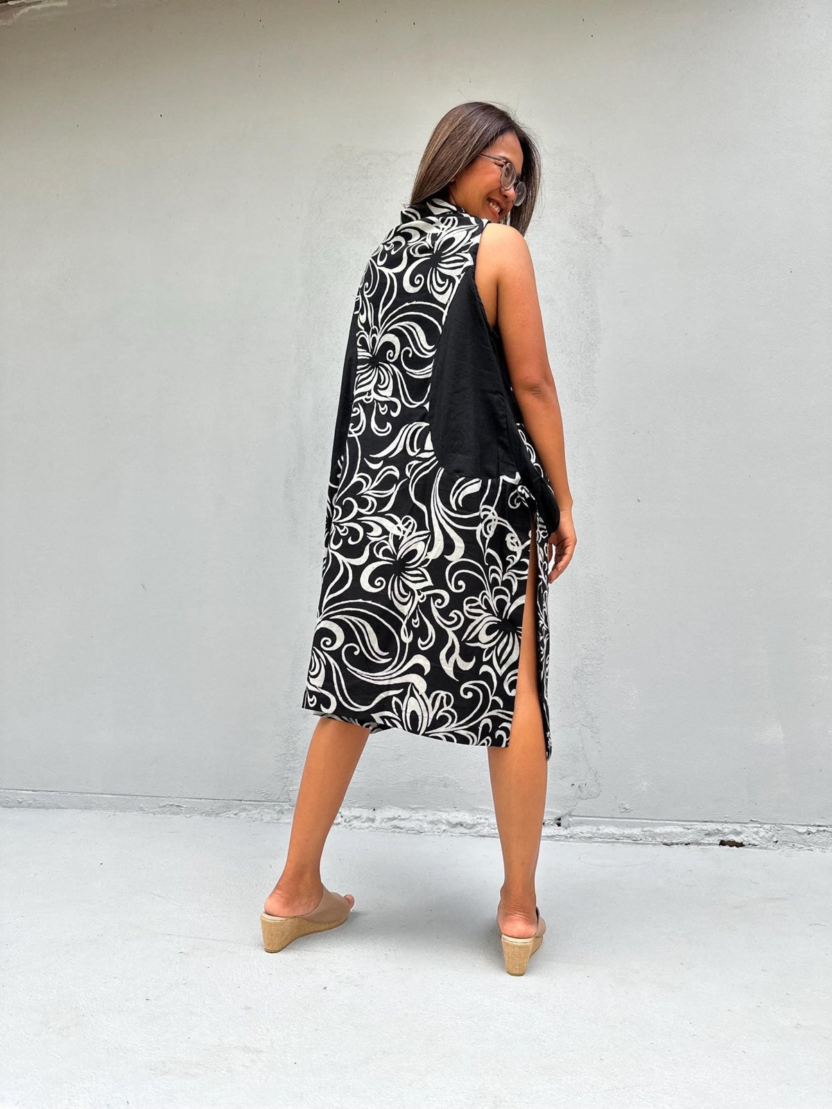 A woman wearing a sleeveless black and white patterned midi dress with a high collar, photographed from behind against a plain light gray wall.