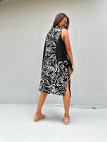 A woman wearing a sleeveless black and white patterned midi dress with a high collar, photographed from behind against a plain light gray wall.