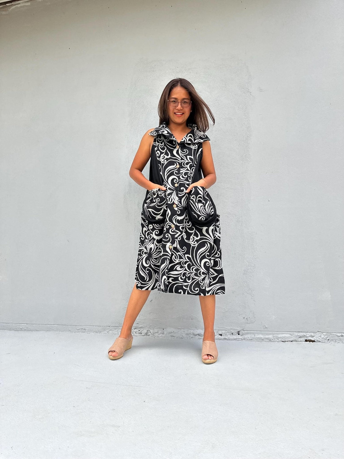A woman modeling a sleeveless midi dress with a black and white swirling damask pattern, standing against a light gray concrete wall.
