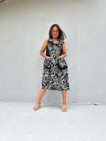 A woman modeling a sleeveless midi dress with a black and white swirling damask pattern, standing against a light gray concrete wall.